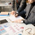 Confident businessman sitting in wheelchair at his workplace and signing a contract with his partner standing near by him